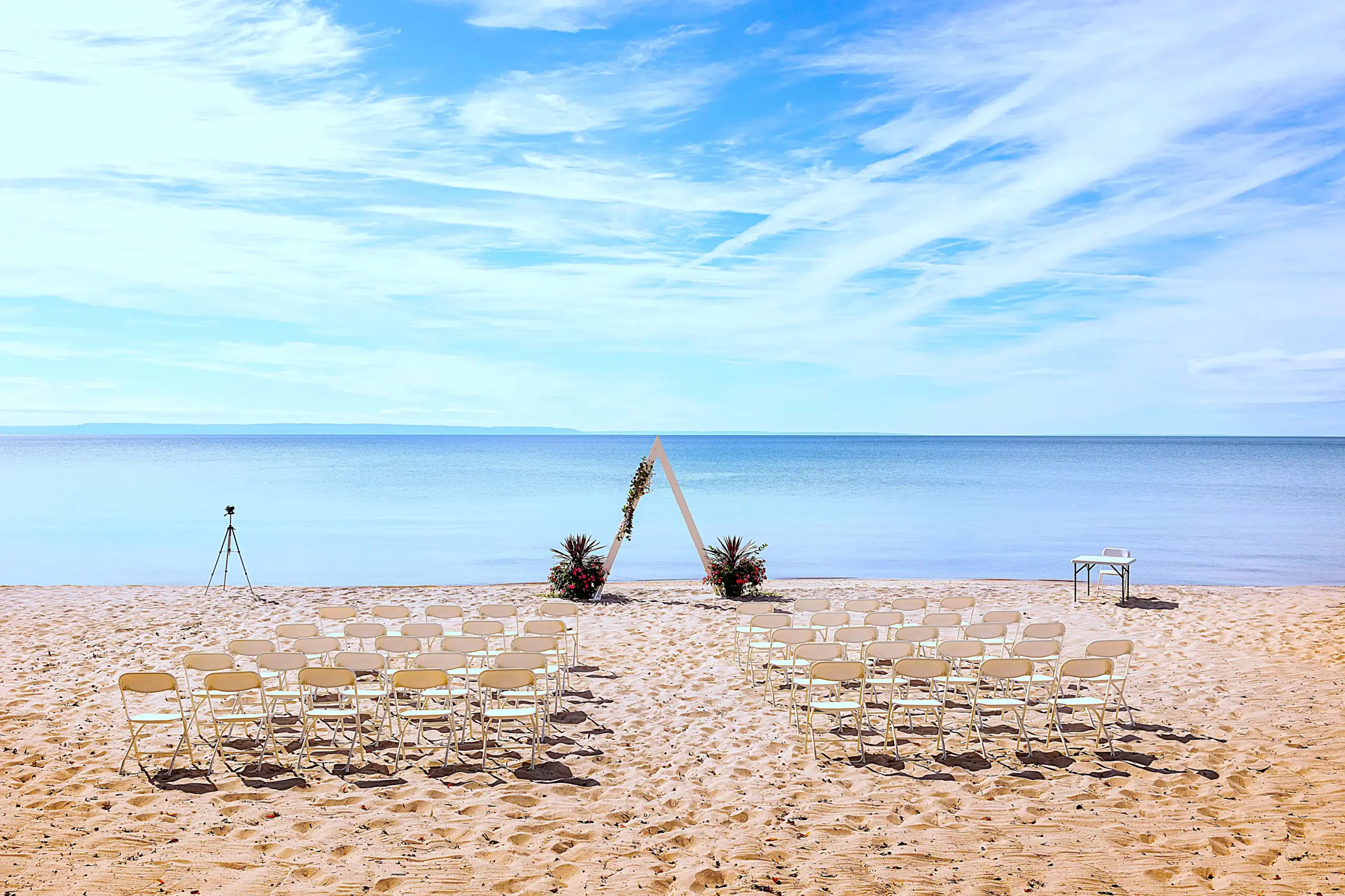 CARI beach wedding ceremony setup in Tiny, Ontario, with white chairs, an A-frame arch, and clear blue water background.